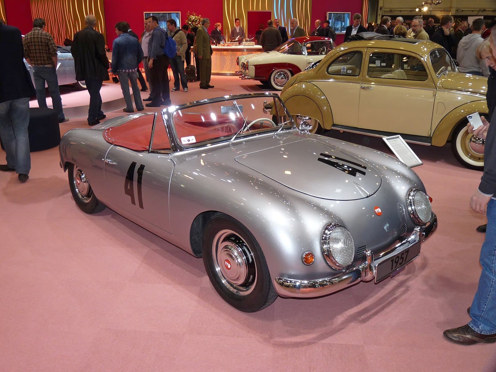 Ein silberner Denzel-Sportwagen von 1957 mit Startnummer auf einer Messe, dahinter ein Käfer