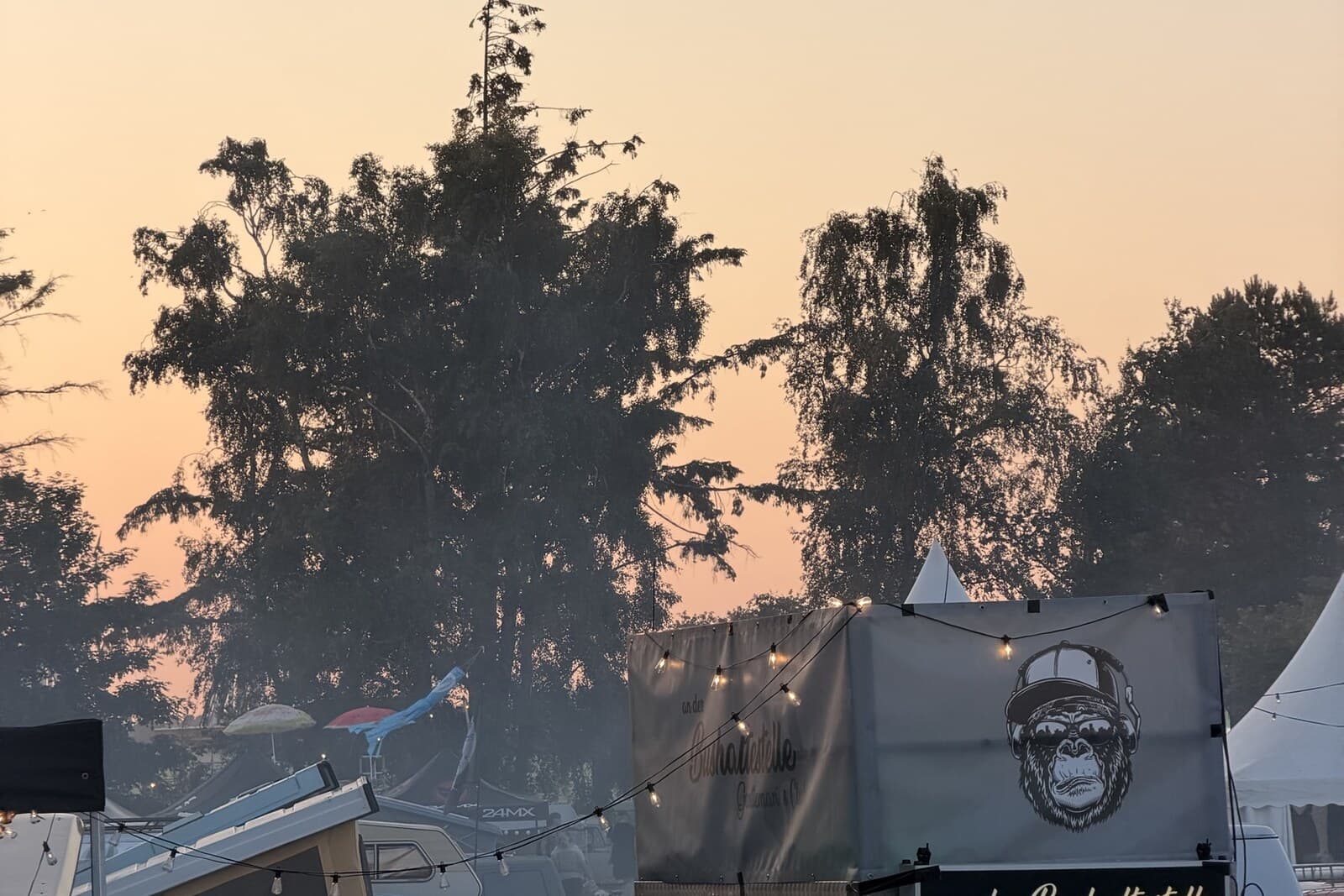 Wenn die Sonne runtergeht, verschiebt sich das Festival an die Bars und Lichterketten. Eigene Aufnahme vom Festival. Abendhimmel über dem Festivalgelände, Lichterketten an einer Bar, Aufstelldächer dahinter