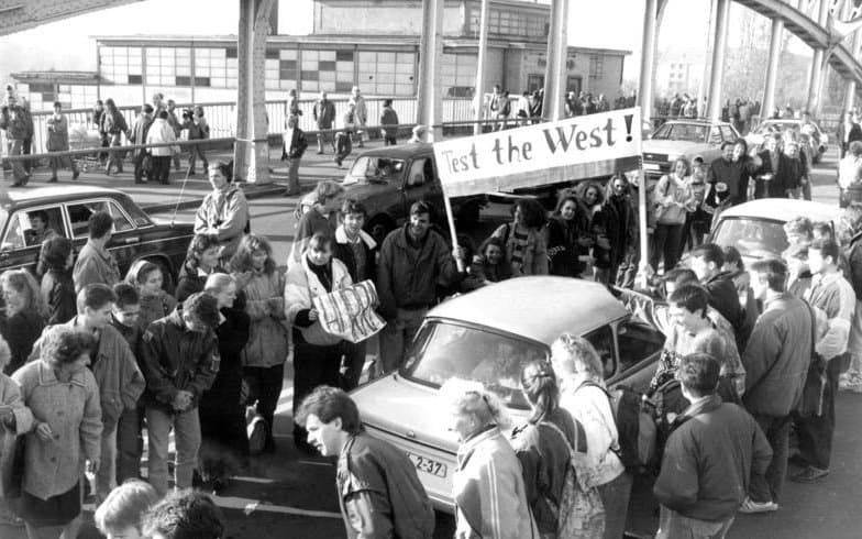 Bornholmer Straße, 10. November 1989: Schüler aus dem Berliner Wedding begrüßen die ersten Autos aus dem Osten. Foto: Hans Peter Lochmann, Bundesarchiv, Bild 183-1989-1118-028, CC BY-SA 3.0 de. Schüler stehen an der Bornholmer Straße Spalier und begrüßen einen Trabant, dahinter ein Plakat mit der Aufschrift Test the West