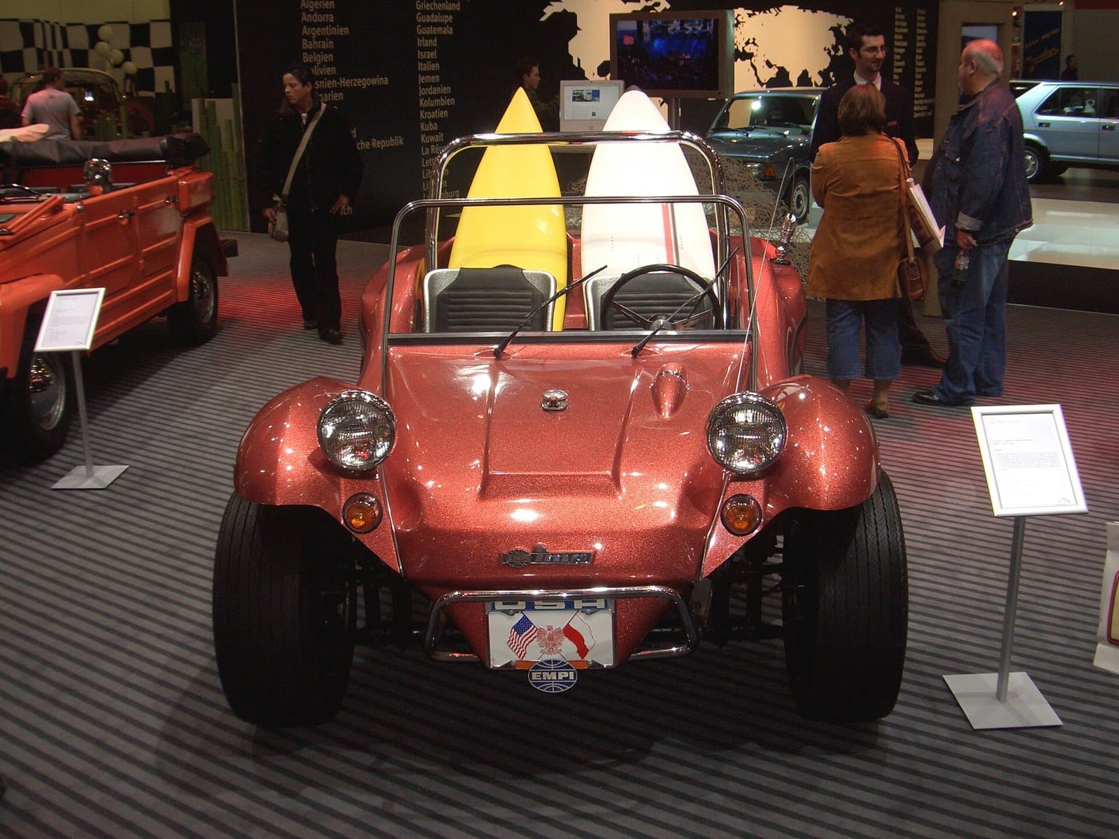 Ein EMPI Imp von 1968, ein Buggy auf Käfer-Basis, mit der EMPI-Plakette vorn. Foto: Detectandpreserve, Wikimedia Commons, CC BY-SA 3.0, skaliert. Ein orangefarbener EMPI Imp Buggy von 1968 mit Surfbrettern auf einer Ausstellung