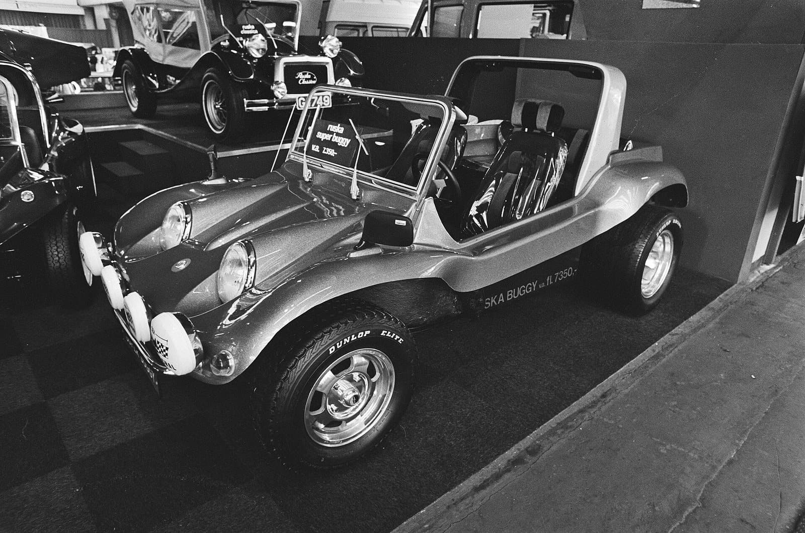 Ein Ruska Super Buggy auf der Automesse RAI in Amsterdam, 4. Februar 1981. Als Super Standaard für 7.350 Gulden war er das günstigste Auto der Messe. Foto: Rob Bogaerts, Anefo, Nationaal Archief, Wikimedia Commons, CC0, skaliert. Schwarzweißfoto eines Ruska Super Buggy mit Preisschild auf einem Messestand